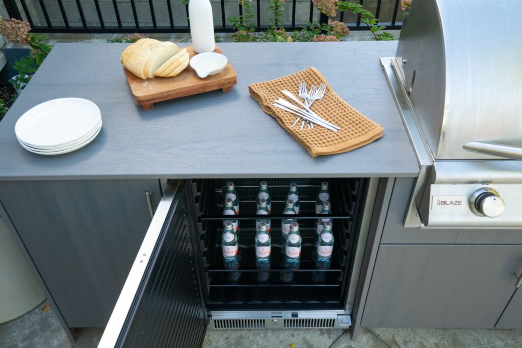 Gray Agora outdoor cabinet beverage fridge open with San Pellegrino bottles, sliced bread and forks staged on top.