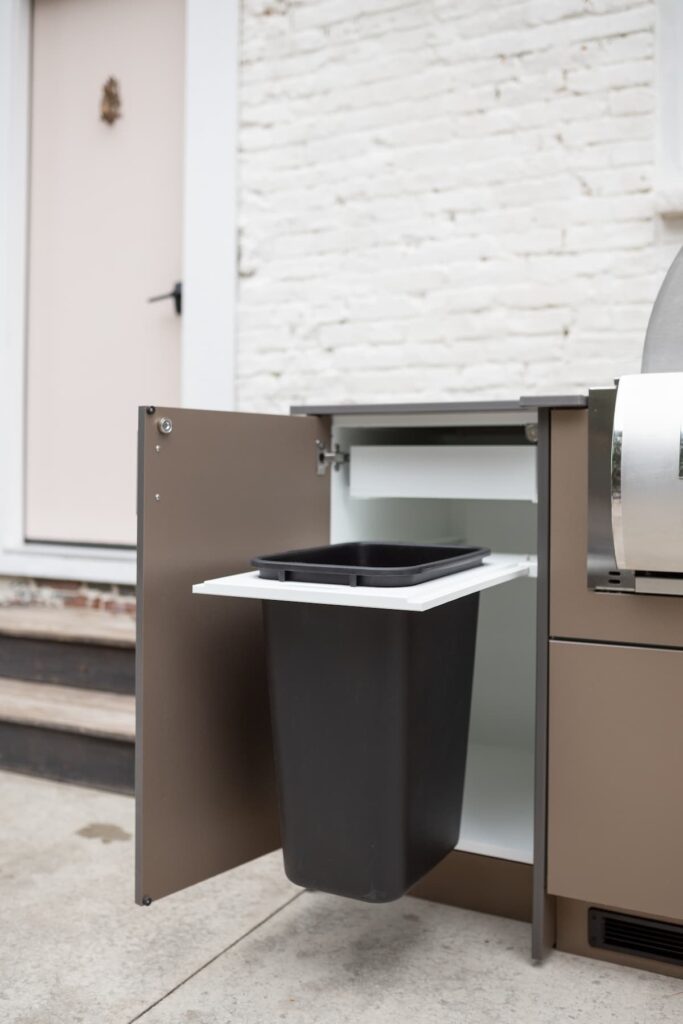Taupe Agora outdoor cabinet with the pull-out trash bin door swung open, exposing the black waste bin beside a grill.