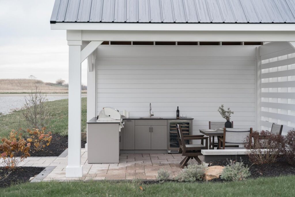 Gray Agora outdoor kitchen with grill, sink, and wine fridge under a white pavilion overlooking a pond and dining set.