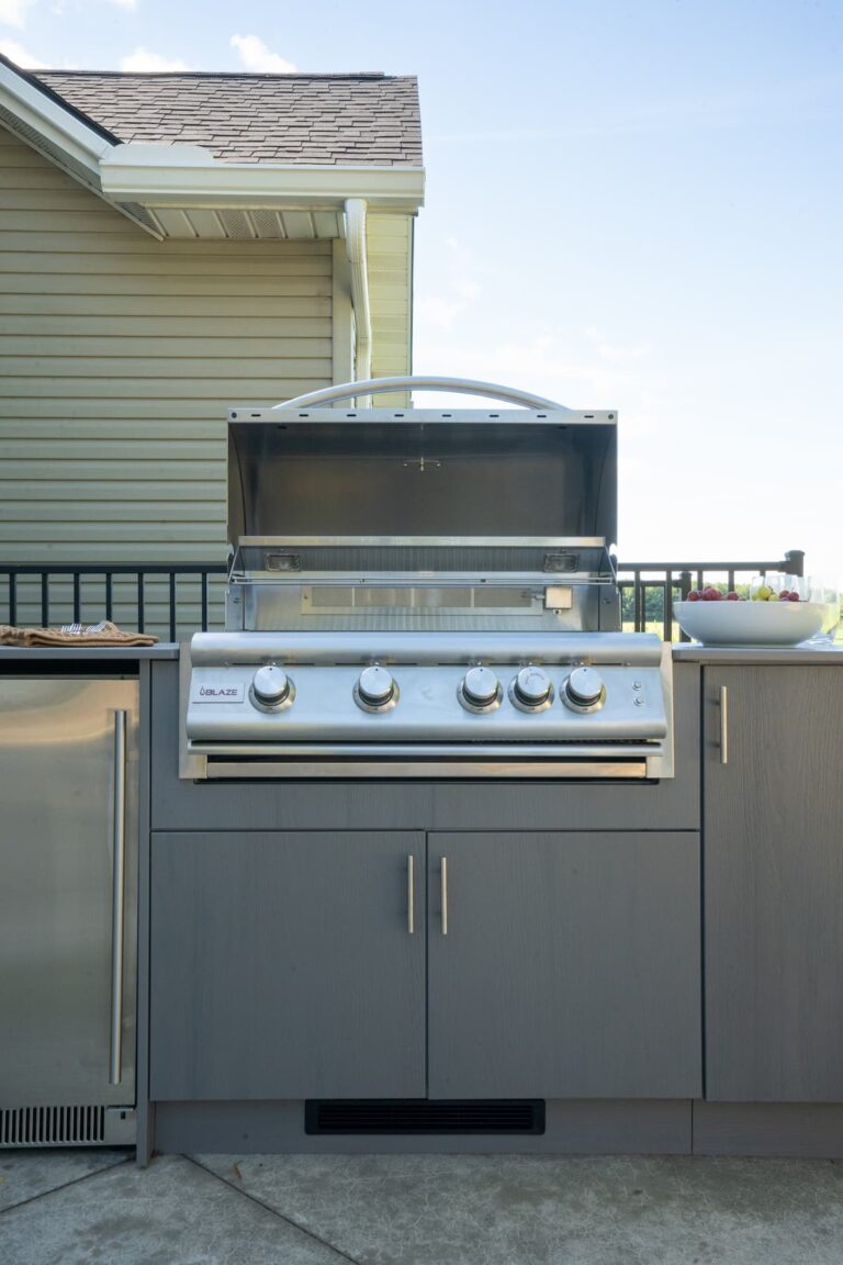 Blaze built-in gas grill with hood open in a gray Agora outdoor cabinet beside a stainless fridge and bowl of grapes.