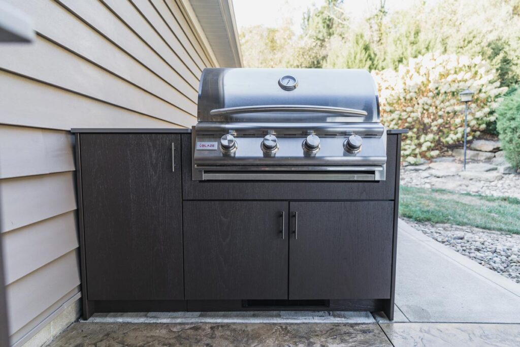 Espresso Agora outdoor cabinet housing a Blaze stainless gas grill with the lid closed against tan siding on a patio.
