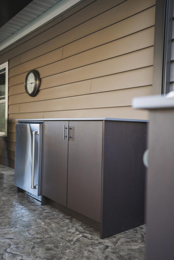 Espresso Agora outdoor cabinets with a built-in stainless refrigerator beneath a wall clock on tan lap siding.