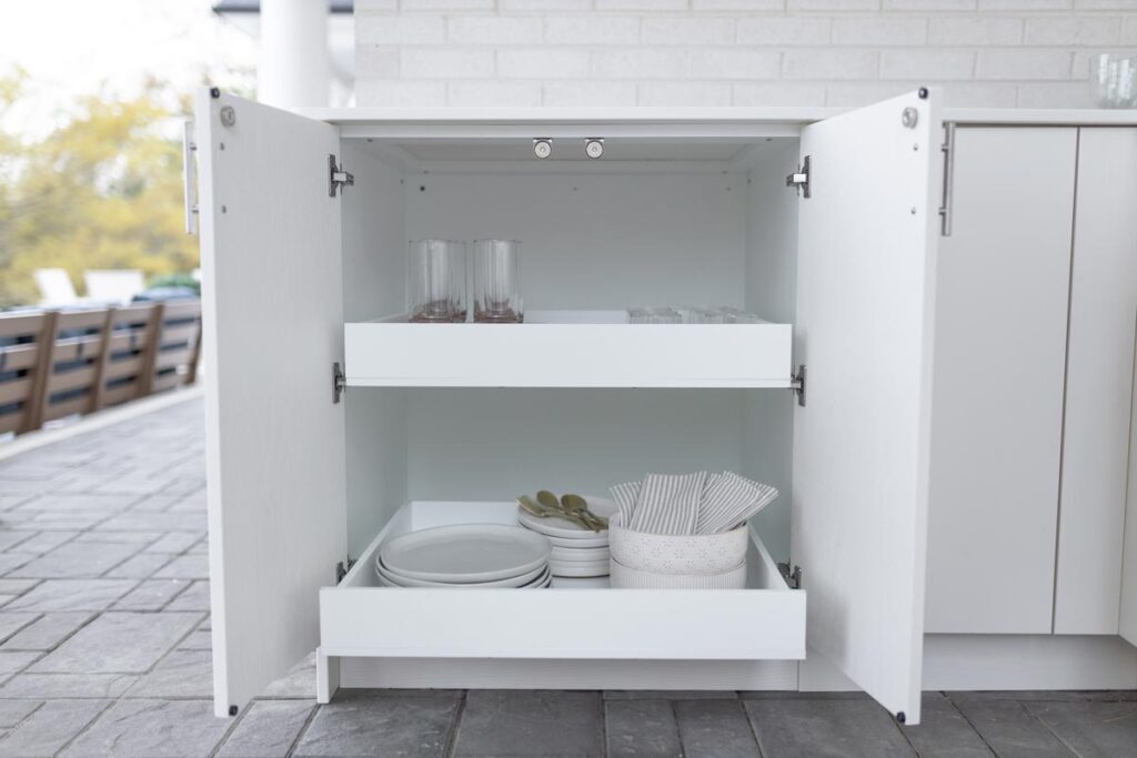 White Agora outdoor cabinet with both doors open showing two pull-out drawers stocked with glasses, plates, and napkins.
